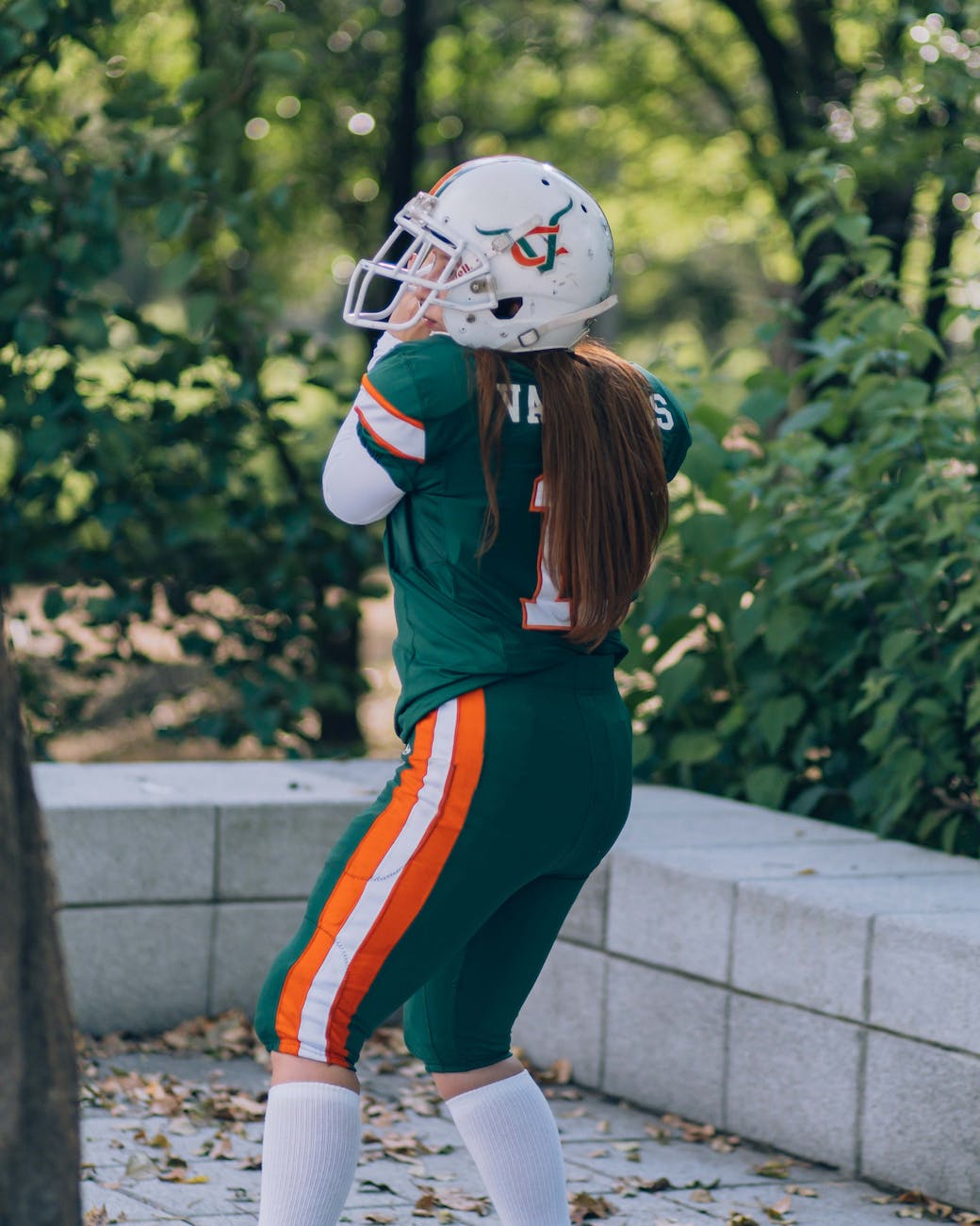 young and pretty millennial athletic girl dressed in a green american football uniform with orange and a background full of plants and trees